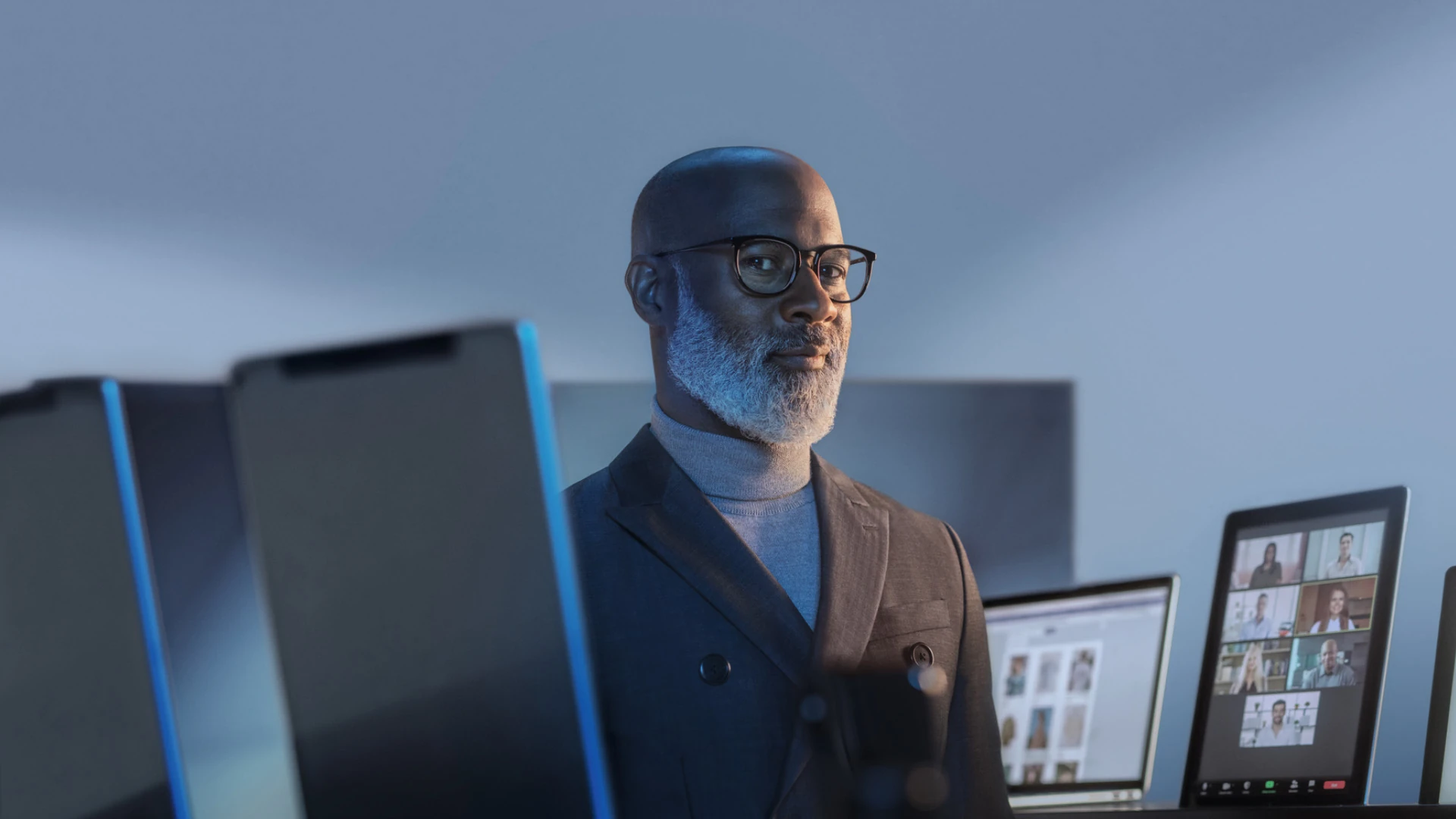 Homem com óculos de lentes incolores entre monitores em um ambiente de trabalho.